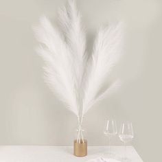 two wine glasses and a vase with white feathers in it sitting on a table next to an empty glass