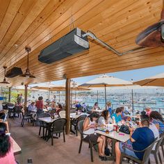 people are sitting at tables with umbrellas over them and boats in the water behind them