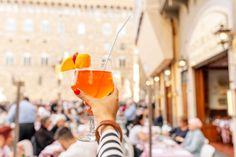 a person holding up a drink in front of an outdoor restaurant with people sitting at tables