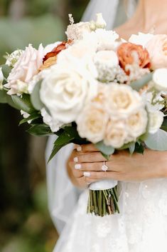 a bride holding a bouquet of white and peach flowers in her hands while wearing a wedding ring