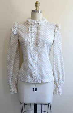 "This precious floral blouse epitomizes the iconic '80s-does-Edwardian fashion trend. Crisp white lace accentuates the shoulders, outlines the high ruff-like collar, and trims the button placket and cuffs. The smooth woven fabric features a pattern of pale pink flowers and blue-green vines against a cream- or ivory-colored backdrop. The blousy sleeves complete the look. Buttoned front closure. Superb vintage condition. No holes, picks, or other significant damage that we could spot. We removed t Spring Retro Blouse With Ruffles, Retro Ruffled Blouse For Spring, Feminine Vintage Fashion Blouse, Feminine Blouse For Vintage Fashion, Fall Vintage Blouse With Vintage Pattern, Vintage Fitted Top With Ruffles, Vintage Blouse With Vintage Pattern For Fall, Fitted Vintage Top With Ruffles, Vintage Fashion Tops For Spring