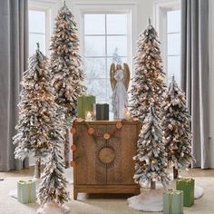 christmas trees in front of a window with presents on the floor and an angel statue next to them