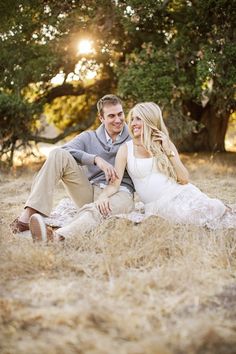 a man and woman are sitting on the ground in front of trees, smiling at each other