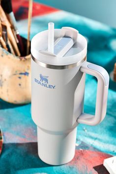 a white travel mug sitting on top of a table next to paintbrushes and brushes