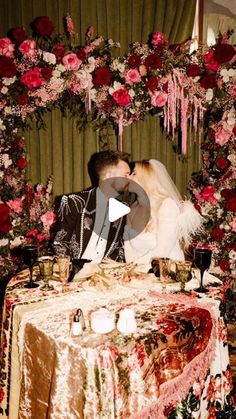 a bride and groom kissing in front of a table with red flowers on the wall