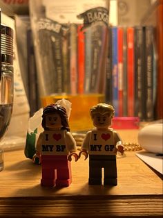 two lego people standing next to each other on a wooden table with books in the background