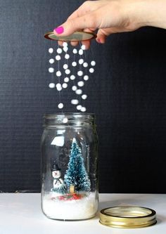 a hand is sprinkling snow into a glass jar filled with trees and snowballs