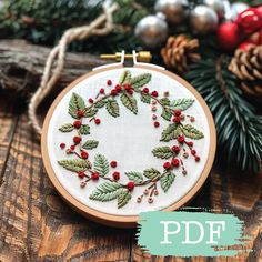 a cross stitch christmas wreath on a wooden table with pine cones and ornaments in the background