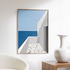 a white vase sitting on top of a wooden table next to a wall mounted painting