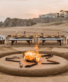 a fire pit on the beach with tables and chairs set up around it for an outdoor dinner