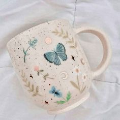 a white coffee cup with blue butterflies and flowers on it's side sitting on a bed