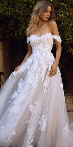 a woman wearing a white wedding dress with flowers on the skirt and off - shoulder