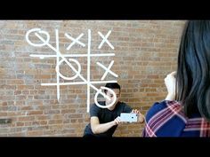 a woman taking a photo of a man using a cell phone in front of a tic - tac - toe wall