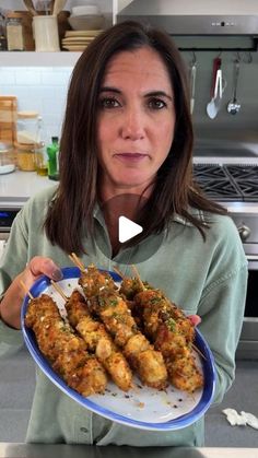 a woman holding a plate with chicken skewers on it in the middle of a kitchen