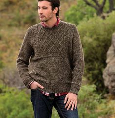 a man standing in the grass wearing a brown sweater and blue jeans with his hands in his pockets