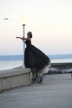 a woman in a black dress leaning against a light post near the ocean at sunset