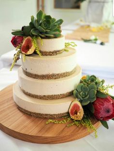 a three tiered cake with flowers and succulents sits on a wooden platter