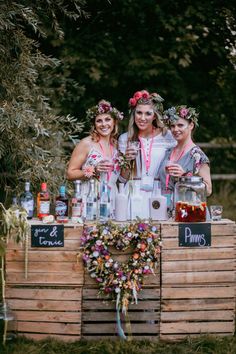 three women standing behind a wooden cart with drinks on it and flowers in their hair