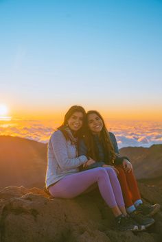 two women sitting on top of a mountain at sunset