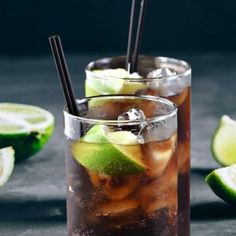 two glasses filled with ice and limes on top of a black table next to sliced limes