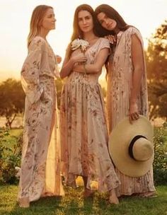 three women in long dresses and hats standing on the grass with their arms around each other