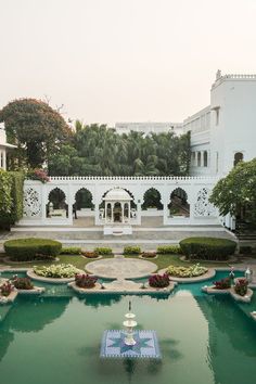 an outdoor fountain surrounded by greenery in front of a white building with columns and arches