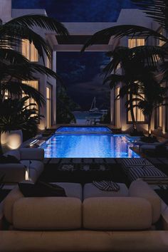 a living room filled with furniture next to a large swimming pool at night in front of tall windows