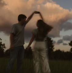 a man and woman dancing in a field under a cloudy sky with their hands together