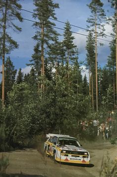 a rally car driving down a dirt road in front of some tall trees and people