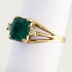 "This magnificent vintage ring boasts a lovely emerald and two diamonds set in 18K yellow gold. The emerald has a rich bluish-green color and weighs in at 1 1/4 carats. An accent diamond is set on each side of the emerald, adding elegance and sparkle. The ring has triple branched shoulders that add interest beyond the classic shank. Luxurious! Vintage: 1990s Size: 6 Materials: 18K Yellow Gold, Emerald, Diamonds Marked: 18K Emerald: 7 mm x 7mm x 4.4mm, approx. 1.25 ct. 2 Diamonds: brilliant cut, Timeless Gold Diamond Ring With Emerald, Formal Emerald Three-stone Ring, Timeless Emerald Diamond Ring In Yellow Gold, Vintage Green Emerald Ring With Brilliant Cut, Timeless Yellow Gold Emerald Diamond Ring, Timeless Yellow Gold Diamond Ring With Emerald, Timeless Green Diamond Ring With Accents, Classic Yellow Gold Emerald Wedding Jewelry, Classic Green Emerald Ring With Diamond Accents