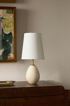 a white lamp sitting on top of a wooden dresser next to a framed flower painting