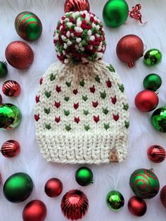 a white knitted hat with red and green pom - poms surrounded by christmas ornaments