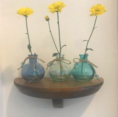 three glass vases with yellow flowers in them on a wooden shelf against a white wall