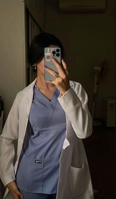 a woman in scrubs taking a selfie with her cell phone while wearing a white lab coat