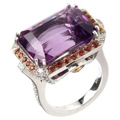 A splendid ring 18kt white gold showcases a mesmerizing center emerald-cut amethyst, which exudes an air of regal elegance. Encircling this captivating amethyst are vibrant orange round sapphires, creating a striking contrast that adds a touch of warmth and drama. To complete this exquisite composition, diamonds adorn the sides of the ring, adding a brilliant sparkle that complements the gemstones. Ring Emerald Cut Amethyst 12.52 cts. with a Halo of 24 Orange Sapphires 1.28 cts., Diamonds 0.92 cts., and 2 trillion Sapphires 0.11 cts. in 18kt White Gold. Size 5.75 Perfect gift for Christmas, Mom, Girlfriend, Daughter, Graduation, Birthday and more. Luxury White Gold Amethyst Ring, Luxury Purple Gemstones With Halo Setting, Formal Purple Sapphire Ring With Gemstone Accents, Luxury White Gold Amethyst Ring With Diamond, Luxury Purple Platinum Rings, Luxury Amethyst Ring With Emerald Cut Diamond, Luxury White Gold Amethyst Ring With Brilliant Cut, Luxury Emerald-cut Diamond Amethyst Ring, Luxury White Gold Amethyst Ring With Gemstone Accents