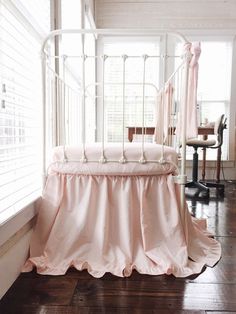 a white crib with pink ruffled bedding in a room next to a window