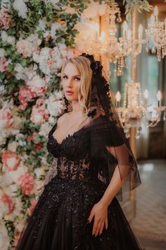 a woman in a black wedding dress standing next to a flower covered wall with chandeliers