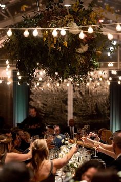 a group of people sitting around a dinner table with wine glasses on it and lights hanging from the ceiling