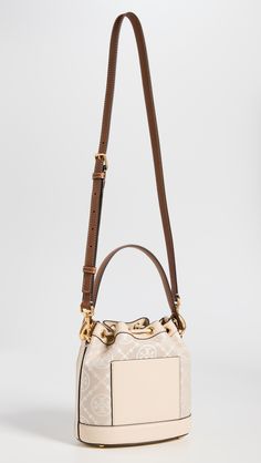 Find TORY BURCH T Monogram Bucket Bag on Editorialist. This Tory Burch bucket bag features a structured silhouette with gold-tone hardware. The bag is crafted from jacquard with a monogram pattern and leather trim. It has a drawstring top, patch interior pocket, and an optional adjustable strap. The bag is lined in suede and has a height of 8.25 inches, a length of 8.25 inches, and a depth of 4.75 inches. The strap drop is 21.75 inches and the handle drop is 4 inches. Luxury Bucket Bag Tote With Gold-tone Hardware, Luxury Bucket Bag With Gold-tone Hardware, Elegant Bucket Bag With Brass Hardware, Elegant Top Handle Bucket Bag With Brass Hardware, Beige Monogram Canvas Bag With Gold-tone Hardware, Luxury Bucket Bag With Brass Hardware, Designer Cream Bucket Bag, Designer Cream Bucket Bag With Detachable Handle, Gold Monogram Canvas Shoulder Bag With Double Handle