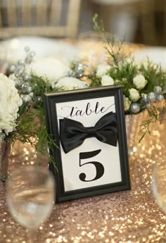 the table numbers are decorated with white flowers and greenery, along with a black bow tie