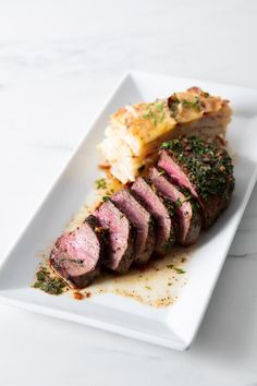 a white plate topped with meat and potatoes on top of a marble countertop next to a knife