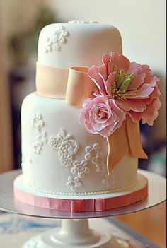 a three tiered wedding cake with pink flowers and ribbons on the top, sitting on a table