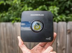 a hand holding up a small black device in front of a fence with trees behind it