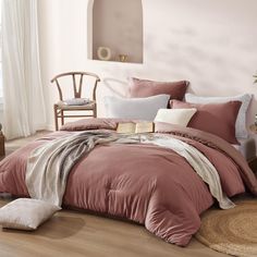 a bed with pink comforter and pillows in a room next to a chair, table and window