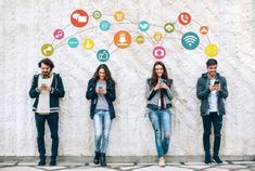 four people are standing in front of a wall with social media icons on it and looking at their phones