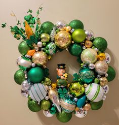 a wreath with green and gold ornaments hanging on the wall next to a white wall