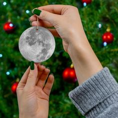 two hands holding an ornament with the moon on it in front of a christmas tree