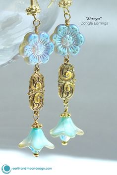 These striking dangle earrings are full of alluring color and shimmer. Lovely bell flowers in dreamy aqua-blue hue with a delicate gold finish and are made of Czech glass and donned with gold-toned pewter embellishments and finished with an aqua-blue Swarovski crystal. They are suspended from decorative gold-toned pewter accents and Czech glass daisies that are washed with an iridescent purple-blue-gold finish. Earrings fall 2" from ornate gold-plated brass ear wires. $36 Feminine Drop Earrings For Spring, Feminine Spring Dangle Earrings, Spring Feminine Dangle Jewelry, Feminine Spring Dangle Jewelry, Delicate Dangle Earrings For Spring, Spring Party Jewelry With Matching Earrings, Elegant Spring Chandelier Earrings, Spring Wedding Drop Earrings, Handmade Feminine Dangle Flower Earrings