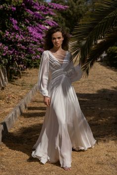 a woman in a long white dress walking down a path with purple flowers behind her