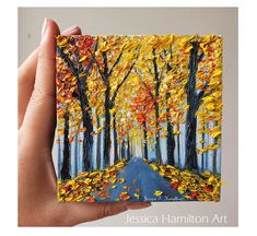 a hand holding up a painting of trees with yellow and red leaves on the ground